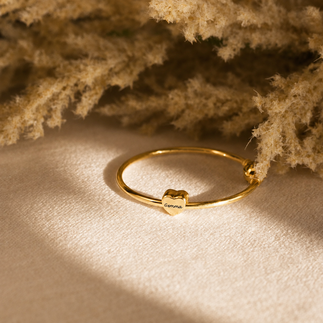 A delicate gold bangle bracelet with a small heart charm engraved with the name 'Gemma', styled on a textured beige surface with dried pampas grass in the background.